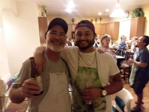 Two smiling men in aprons with arms around each other holding beers, posing at a lively home kitchen party with guests chatting in the background.