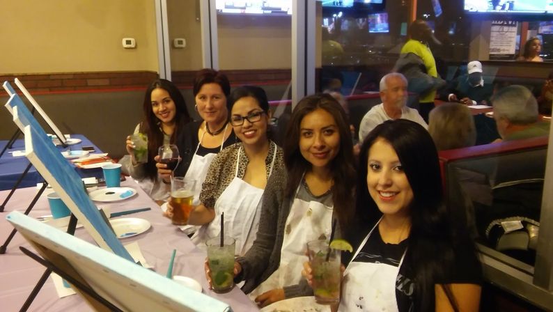 Group of five women in aprons at an indoor paint-and-sip party, seated at easels with canvases and paint supplies, each holding cocktails and smiling in a lively bar-style setting.