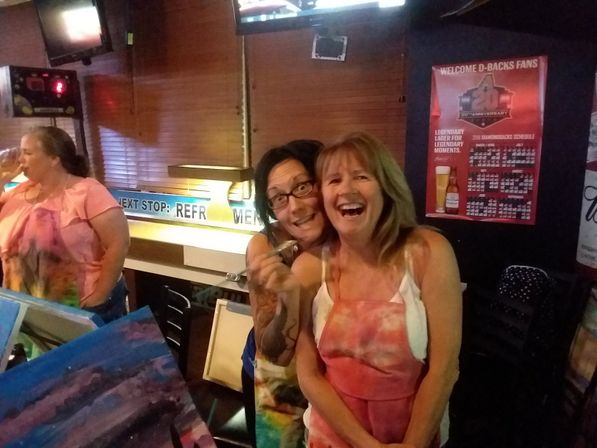 Two laughing women in paint-splattered aprons at a lively paint‑night in a dim bar, holding brushes with canvases and drinks visible.