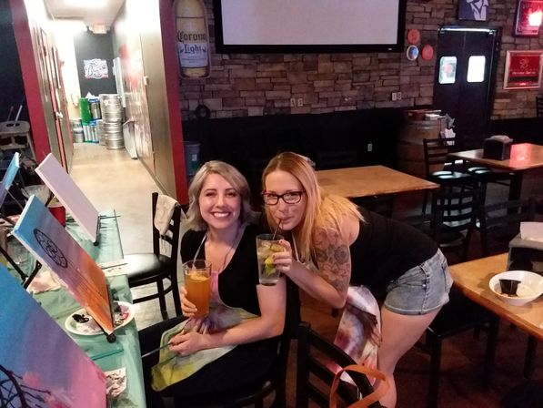 Two smiling women at an indoor paint-and-sip night in a cozy pub-style space, seated at a table with easels, canvases and paint supplies, each holding a cocktail or beer.