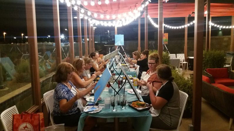 Group of people enjoying a paint-and-sip on an outdoor covered patio at night under string lights, seated along a long table with easels, canvases, drinks and snacks.