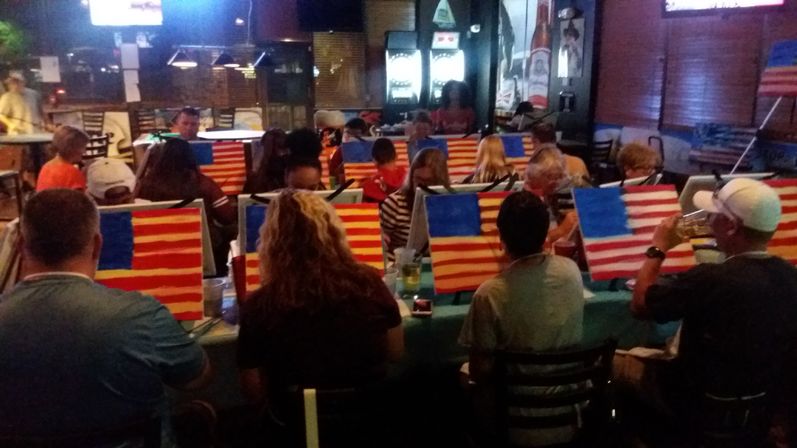 Lively evening paint night in a dim bar with rows of people painting American flag canvases at tables, drinks and art supplies visible.