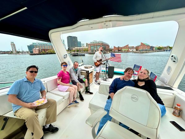 Friends relaxing on a white motorboat in an urban harbor — passengers lounging on cushioned seats while a guitarist performs beside American flags with city skyline across the water.