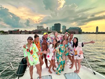 Group of people in colorful summer outfits laughing and posing on the bow of a boat at sunset with an urban waterfront skyline and dramatic clouds in the background.