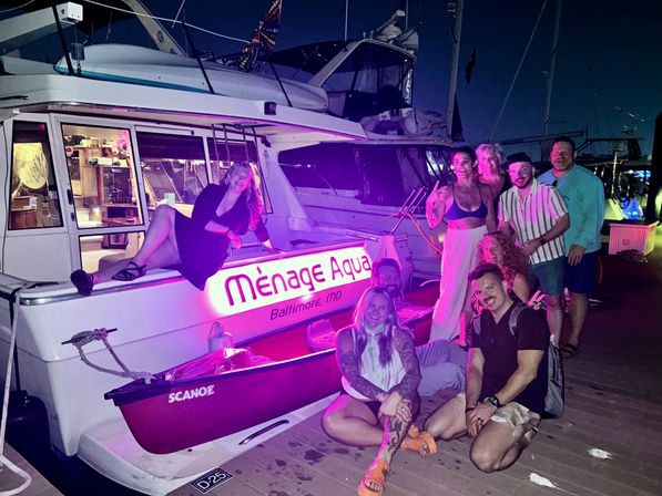 Smiling group posing on and beside a yacht at a marina dock at night, purple neon lighting and a small red canoe attached to the boat.