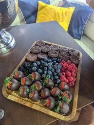 Wooden dessert board with chocolate-covered strawberries, fresh blueberries, raspberries, blackberries and mini chocolate brownies on a dark wood table with colorful throw pillows in the background