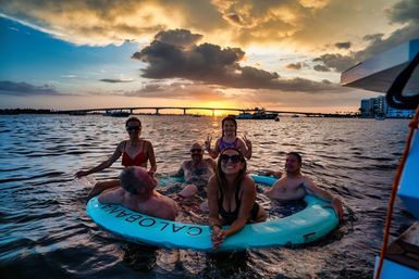 Smiling group of people lounging on a turquoise circular float in a coastal bay at golden sunset, with a bridge, boats and glowing clouds on the horizon.
