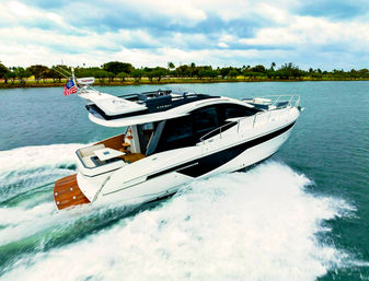White luxury motor yacht speeding along a tree-lined coastal waterway under a cloudy sky, leaving a foamy wake and flying an American flag.