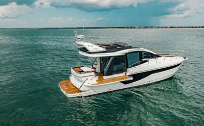 Aerial view of a sleek white luxury motor yacht with teak deck cruising turquoise sea near a distant shoreline under a partly cloudy sky