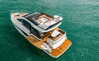 Aerial view of a white luxury motor yacht with teak swim platform, open upper deck seating and helm, floating on clear turquoise sea.