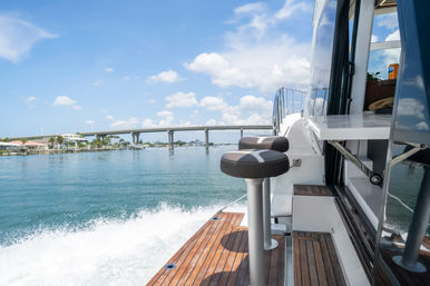 Yacht teak swim platform with two bar stools, wake trailing in turquoise water, coastal bridge and waterfront homes under a sunny blue sky.