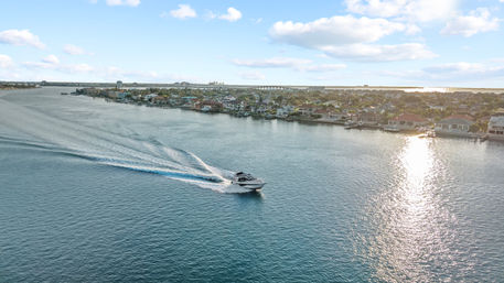 Motorboat speeding across a sunlit coastal inlet, leaving a long wake past waterfront houses and docks with a distant bridge under a blue sky