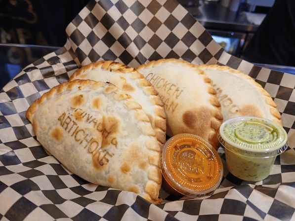 Golden-brown baked empanadas stamped 'ARTICHOKE' and 'CHICKEN' on black-and-white checkered deli paper with two dipping sauces, orange spicy and green herb.