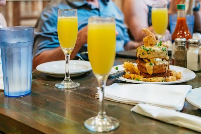 Brunch table with bright orange mimosas in champagne flutes beside a stacked savory breakfast tower topped with crispy fried shrimp and creamy sauce, plus a water glass and condiments.