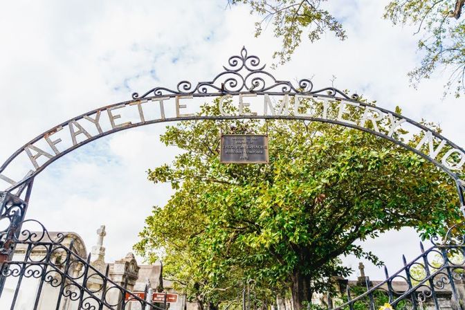 Ornate wrought-iron arch reading "Lafayette Cemetery" over an iron gate, with a large magnolia tree and historic above-ground tombs under a cloudy sky