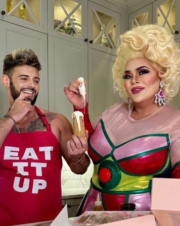 Drag performer in a colorful metallic costume and big blonde wig piping white icing onto a pastry while a smiling man in a red "EAT IT UP" apron watches in a bright home kitchen.
