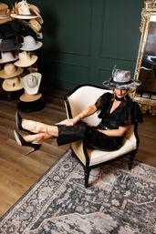 Stylish woman lounging in a cream armchair wearing a feathered fedora and black outfit inside an upscale hat boutique with a hat display rack, ornate gold mirror, green paneled wall and patterned rug