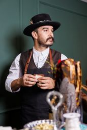 Bearded artisan in a black hat and leather‑strapped apron in a green-walled workshop, holding a small leather patch with craft tools blurred in the foreground.