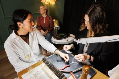 Smiling customer with a glass of champagne getting a bracelet fitted by a jeweler at a cozy boutique jewelry studio, tools and bracelet display on the table and a friend seated in the background.