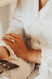 Close-up of hands resting on a white textured dress, featuring a square-cut diamond engagement ring, delicate gold bracelets and a smartphone at the edge in warm tones.