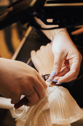 Close-up of hands-on bridal gown alteration: a seamstress with lavender nails fastening a button on a white pleated wedding dress in a studio.