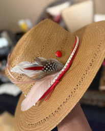 Close-up of a woven straw cowboy hat with wide brim, red ribbon and studded trim, decorative white and speckled feathers and a red accent button — stylish Western summer accessory