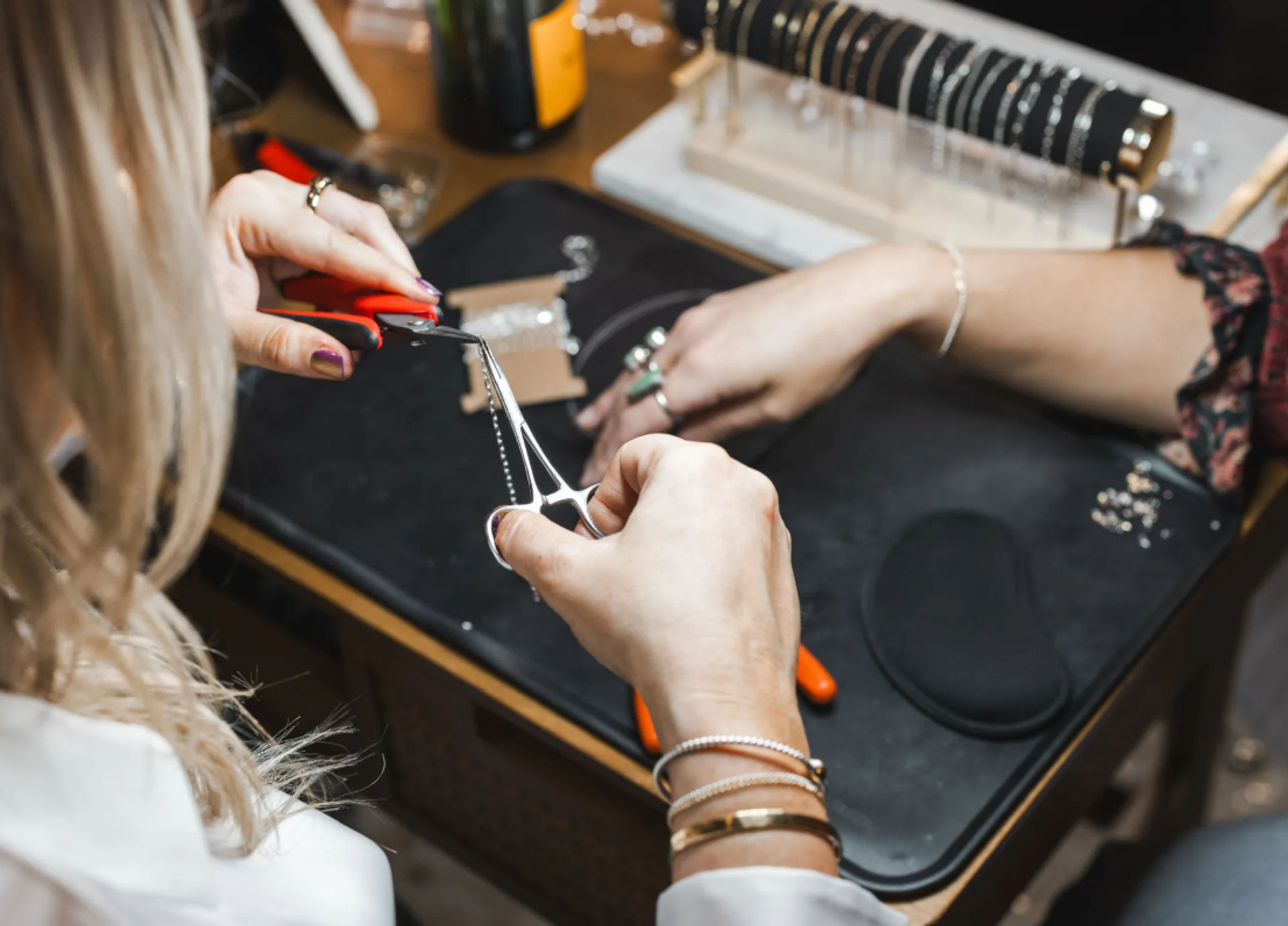 Close-up of hands using red-handled pliers to repair a silver chain necklace on a jewelry workbench, with bracelets and tools nearby