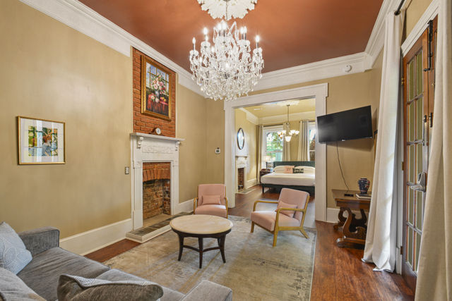 Period-style living room with a sparkling crystal chandelier, exposed brick fireplace with white mantel, blush armchairs and round coffee table, gray sofa on hardwood floors, French doors with curtains and an open view into a sunlit bedroom.