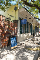 Sunlit sidewalk outside a small boutique hotel in New Orleans with a blue hanging sign and A-frame board by a brick wall, green shutters and dappled oak tree shadows.