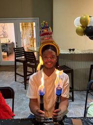 Smiling person at an indoor birthday party wearing a foil cake hat and holding two hand torches with blue-orange flames over a dessert, with black-and-gold balloons and a countertop in the background.