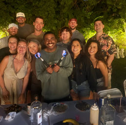 Smiling group of friends posing at a nighttime backyard garden party, man in front holding two lit fire batons above a table with drinks, plates and condiments, tropical foliage and oversized chess pieces in the background