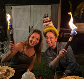 Two smiling women at a backyard birthday party at night — one wearing an inflatable "Happy Birthday" cake hat — both holding lit tiki torches with bright blue flames, dinner plates and a garden shed visible in the background.