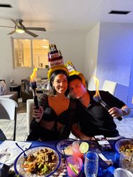 Two smiling people on a backyard patio wearing inflatable 'Happy Birthday' cake hats and holding lit torches over a table with plates of food and colorful cocktails, evening birthday celebration.