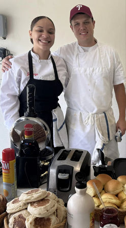 Two smiling chefs in aprons behind a busy breakfast station with a toaster, waffle press, baskets of English muffins and rolls, syrup bottle and jar of jam — cafe kitchen ready for morning service.