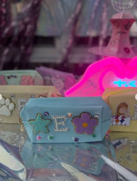 Pastel blue cosmetic pouch decorated with a teal star, beaded letter E, purple flower and gem accents reflected on a shiny surface in a neon-pink boutique window display