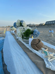 Coastal-chic beach table on sandy shore with burlap runner, white and blue hydrangea centerpieces, baby's breath, a glass candle, nautical rope knot and starfish, oceanfront buildings under a clear sky.