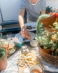 Sunny patio charcuterie spread with cheeses, crackers, cornichons, a tin of sardines, crusty bread and potted herbs while a person in a striped shirt serves from a ramekin