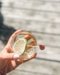 Sunlit close-up of a hand with white nail polish holding a clear cocktail glass garnished with a lime wheel on a skewer topped by a tiny red tomato charm, blurred wooden deck patio background.