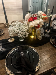 Gothic Halloween tablescape on a rustic wooden dining table: black and gold charger plates with black napkins and skull napkin rings, skull centerpiece with glitter accent, glass vases of white roses, red blooms and baby’s breath, black taper candles and a glowing votive.