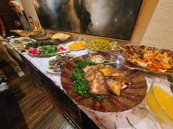 Indoor buffet table with a glittery platter of grilled fish over greens, bowls of roasted vegetables, salads, radishes and a pitcher of orange juice