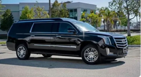 Black luxury SUV with chrome grille and alloy wheels parked in a sunny suburban office-park lot, palm trees and a modern low-rise building in the background.