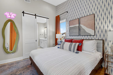 Bright modern bedroom interior with patterned blue-and-white accent wall, queen bed with white textured bedding and red striped pillows, abstract wall art, sliding barn door to en-suite bathroom, green-framed full-length mirror, pink neon sign and orange curtain by window.