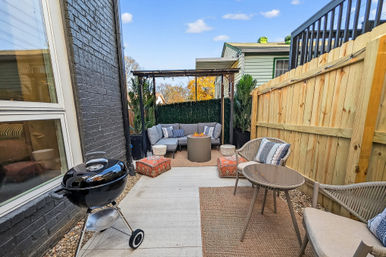 Cozy urban backyard patio with pergola and sectional sofa surrounding a round fire pit, patterned floor cushions, wicker chairs, small charcoal grill by a brick house, wooden privacy fence, potted plants and string lights under a blue sky.