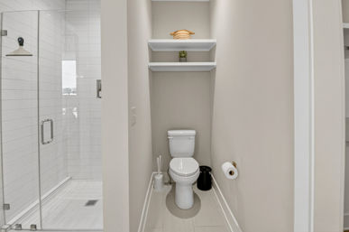 Modern small bathroom water closet in neutral beige: compact white toilet under two floating shelves with succulent and woven basket, wall-mounted toilet paper holder, toilet brush and black waste bin, adjacent glass-enclosed shower with white subway tile.