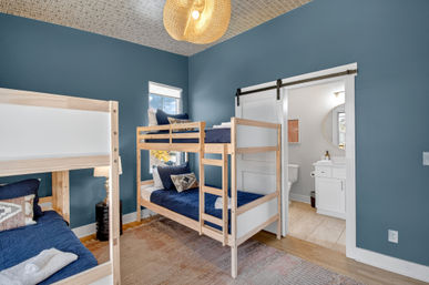Cozy blue-walled bedroom with natural wood twin bunk beds, navy bedding, woven pendant light, area rug, and sliding barn door leading to a bright white bathroom.