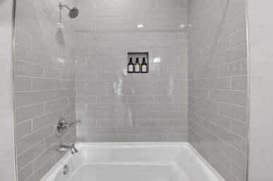 Sleek gray subway-tiled bathtub and shower with white tub, chrome fixtures, and recessed niche holding three shampoo bottles