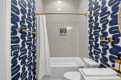 Small modern bathroom with navy blue brushstroke wallpaper, white bathtub and light gray subway-tiled shower, gold-tone fixtures and curved shower rod, recessed niche with amber toiletry bottles, white shower curtain and folded towel