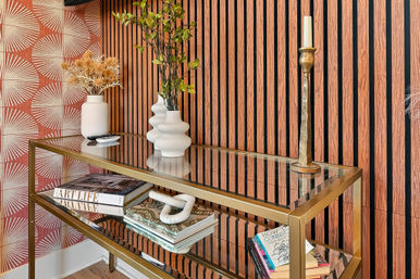 Chic modern entryway with gold-framed glass console table, sculptural white vases, stacked art books, brass candlestick, wood slat accent wall and geometric wallpaper.