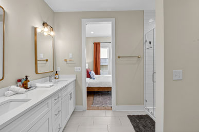 Bright modern ensuite bathroom with white double-sink vanity, gold-framed mirrors and fixtures, glass walk-in shower, neutral tile floor and a view into a bedroom with orange curtains.
