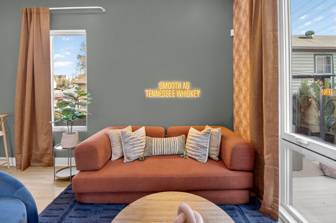 Cozy modern living room with rust-orange sofa and striped throw pillows in front of a gray accent wall featuring a neon sign reading "SMOOTH AS TENNESSEE WHISKEY", tall windows with brown curtains, potted plant, and round wooden coffee table.
